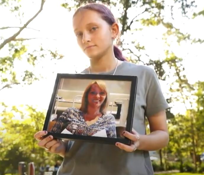 A teenage girl holding a photo framed of her mother standing outdoors with trees behind her